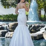 A bride in a long white wedding gown, turned back and striking a pose, standing in front of a small water fountain in an outdoor area surrounded by trees and plants