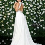 A girl in an ivory white long gown, turned back and striking a pose in front of a wall decorated with white flowers and green plants