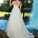 A bride in a long white wedding gown, turned back and striking a pose, standing in front of a small water fountain in an outdoor area surrounded by trees and plants