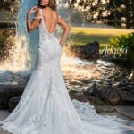 A bride in a long white wedding gown, turned back and striking a pose, standing in front of a small water fountain in an outdoor area surrounded by trees and plants