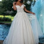 A bride in a long white wedding gown, standing gracefully in front of a small water fountain in an outdoor area, surrounded by trees and plants