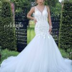 A bride in a long white wedding gown, standing gracefully in front of a small gate. Lush green trees and plants climb a wall behind her, creating a picturesque backdrop