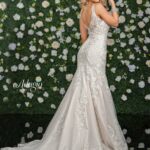 A girl in an ivory white long wedding gown, turned back and striking a pose in front of a wall decorated with white flowers and green plants