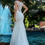 A bride in a long white wedding gown, turned back and striking a pose, standing in front of a small water fountain in an outdoor area surrounded by trees and plants