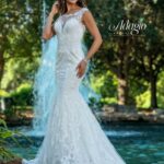 A bride in a long white wedding gown, standing gracefully in front of a small water fountain in an outdoor area, surrounded by trees and plants