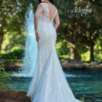 A bride in a long white wedding gown, turned back and striking a pose, standing in front of a small water fountain in an outdoor area surrounded by trees and plants