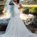 A bride in a long white wedding gown, turned back and striking a pose, standing in front of a small water fountain in an outdoor area surrounded by trees and plants